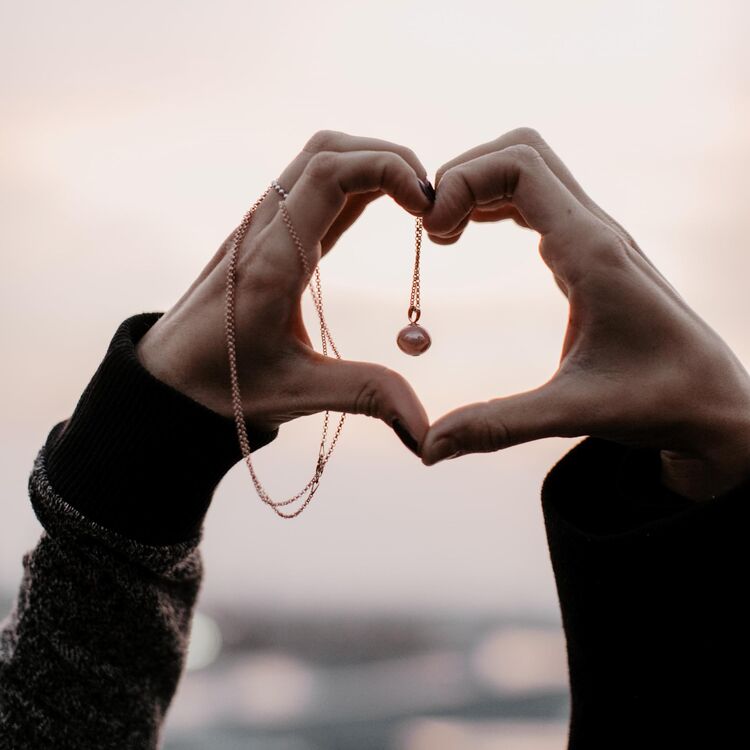 Zwei Hände formen ein Herz. Eine Kette mit einem Anhänger befindet sich zwischen den Händen.