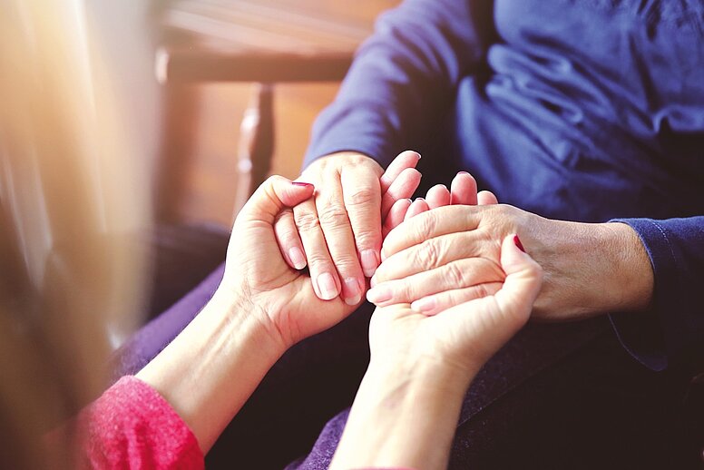 Zwei Personen sitzen sich gegenüber. Die linke Person hält beide Hände ihres Gegenübers in ihren.