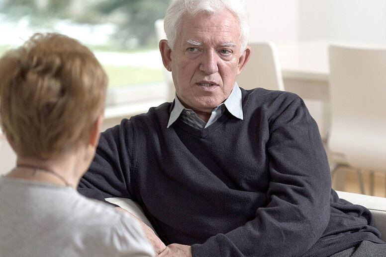 Zwei ältere Personen sitzen sich auf einer Couch gegenüber und unterhalten sich.