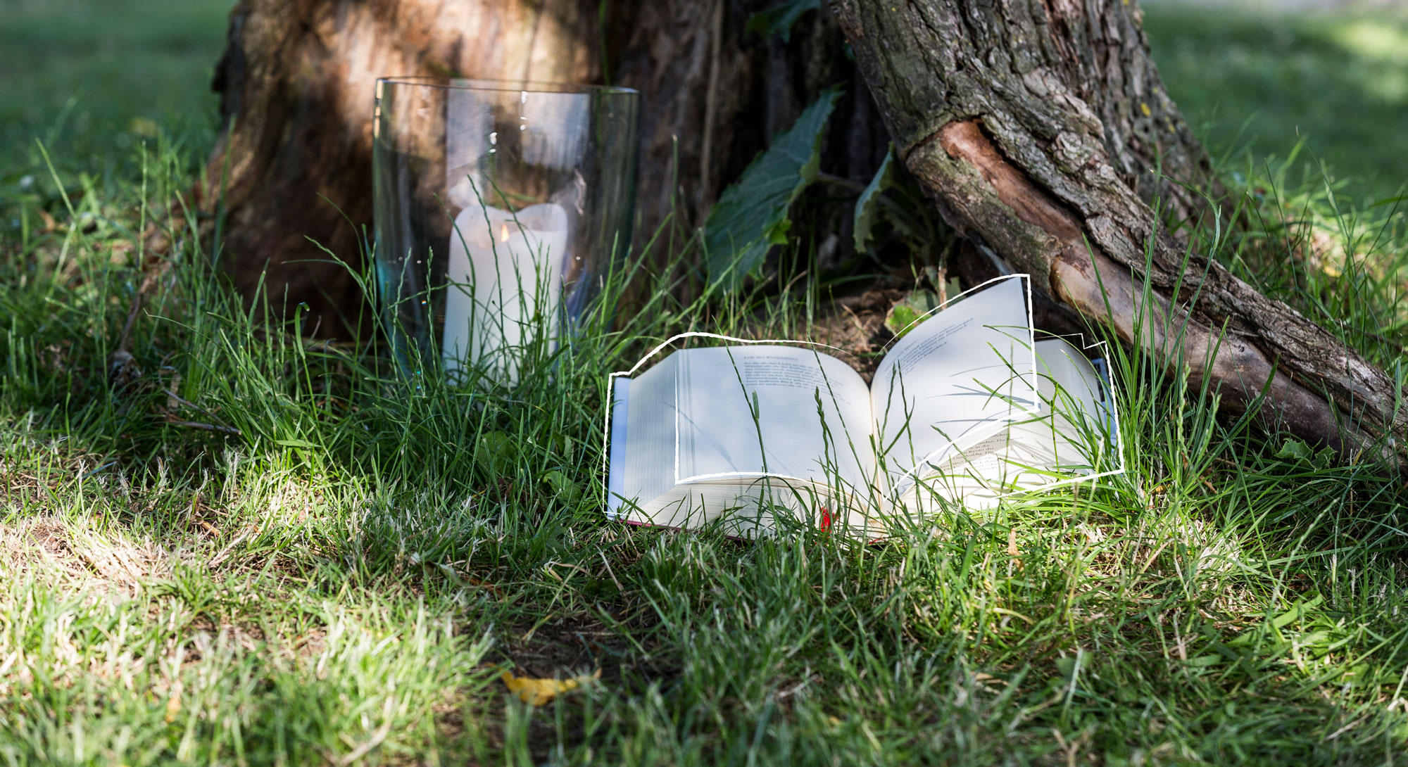 Vor einem Baum liegt ein aufgeschlagenes Buch, links daneben steht eine brennende Kerze in einem Glasgefäß.